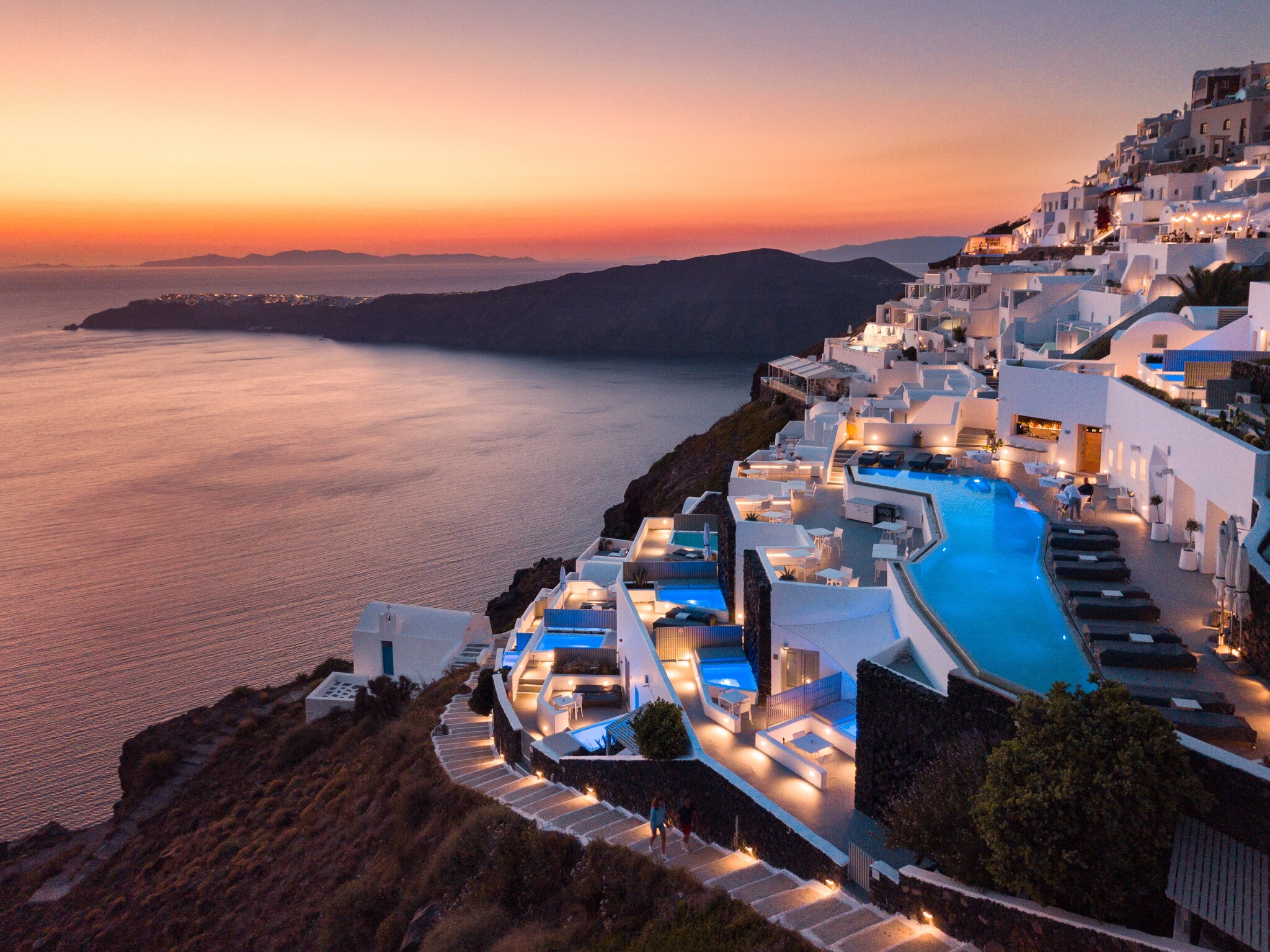 Aerial view of Grace Hotel, Auberge Resorts Collection, at sunset. The white buildings of the Santorini hotel take up a series of terraces.