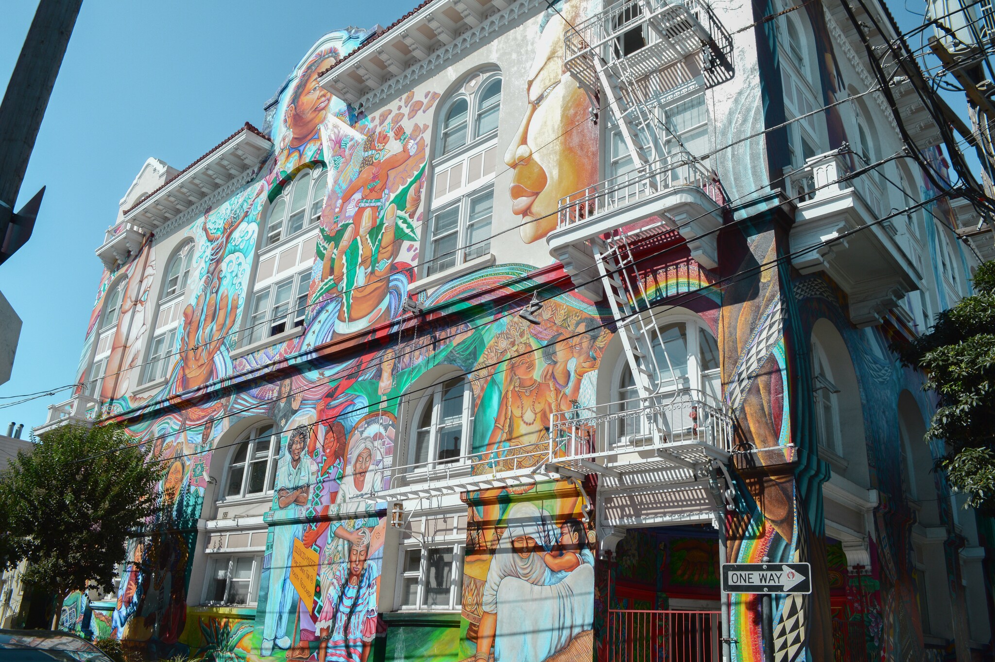 The side of a large white building is covered in a mural, with historical, religious, and mythological figures swirling in rivers of color.