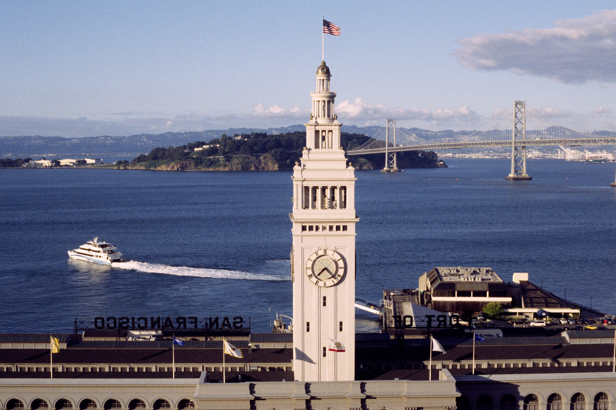 The clock tower of the Ferry Building rises white in front of the bay, with a motorboat and the Oakland Bay Bridge crossing behind.