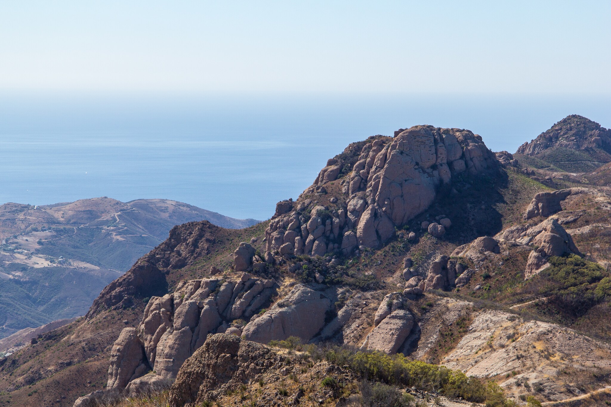 A thin trail winds up a scrubby rock peak. More hills are hazy in the background, with the Pacific ocean beyond.