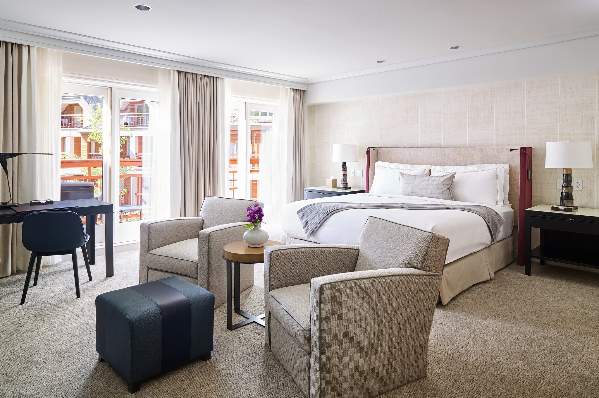 A guest room is furnished with a bed, a sitting area with armchairs and an ottoman, and a black desk. The walls and carpets are light brown.