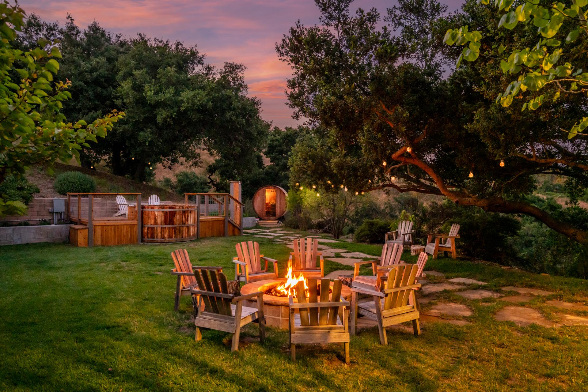 View of outdoor firepit surrounded by Adirondack seating and rustic barrel outdoor hot tub in a grassy area set amongst mature trees at dusk. 