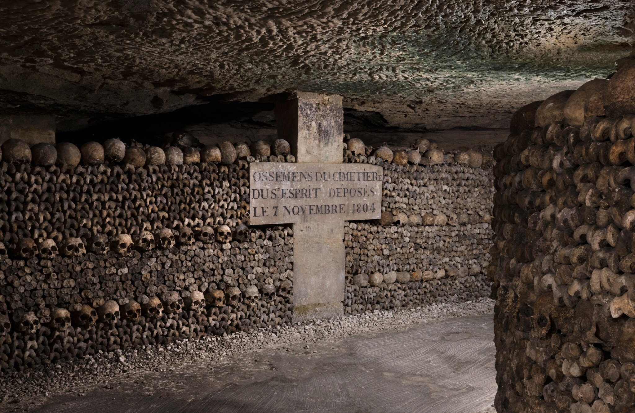 In a hallway of the Paris Catacombs, rows of skulls and bones line the walls, nestled around a large stone cross with carved memorials.