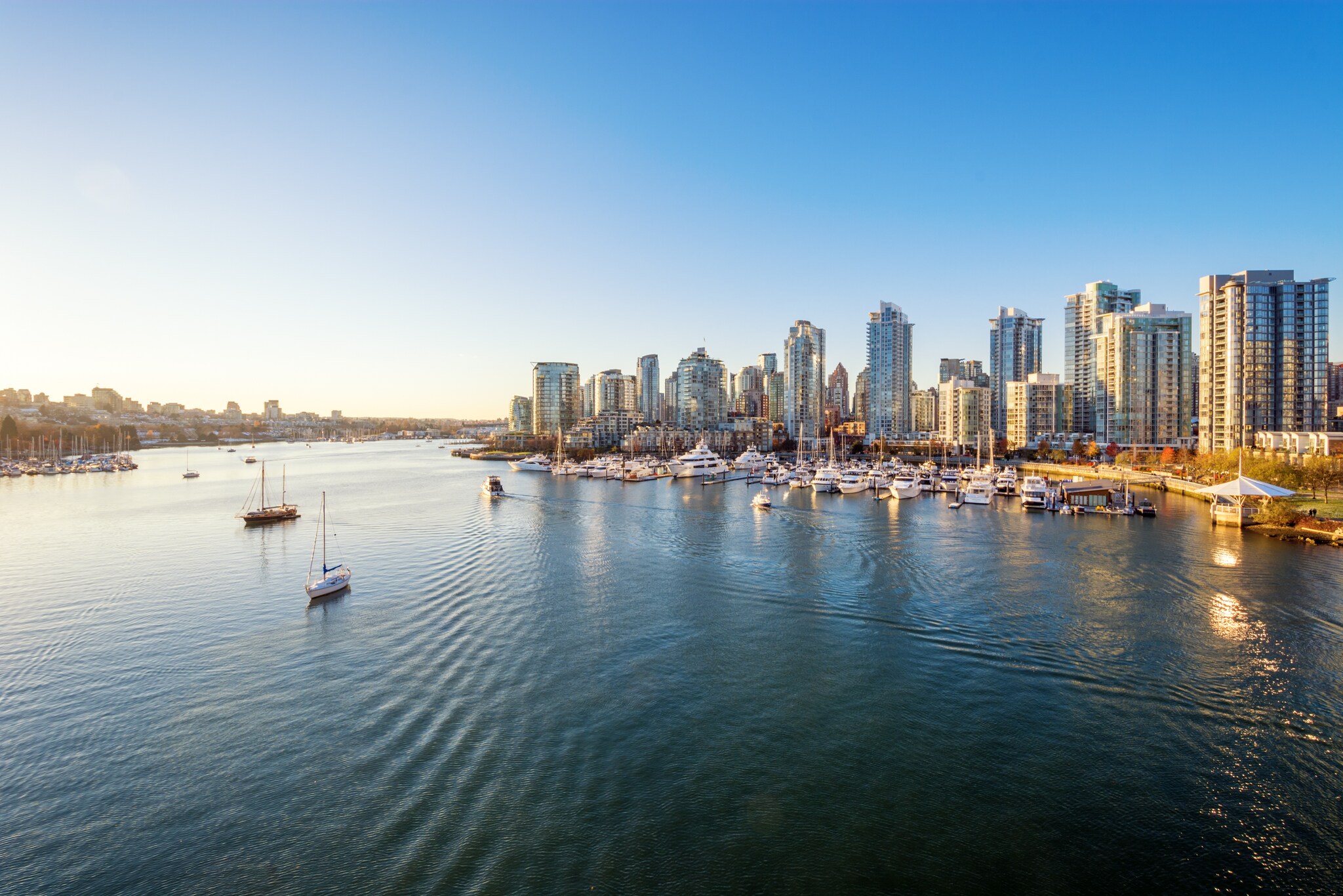Aerial view from the water looking over boats towards the Vancouver skyline, with lines of tall buildings tinged gold by late-day sun.