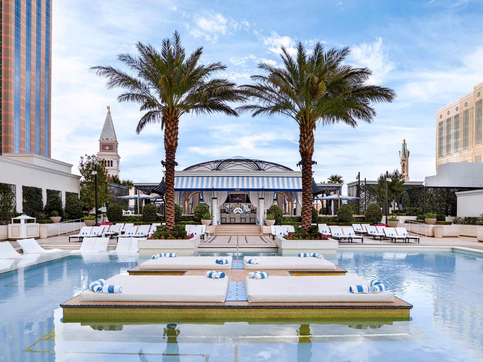 The pool deck at the Venetian Las Vegas boasts a bar framed by palm trees, as well as lounge pads in the center of a large rectangular pool.
