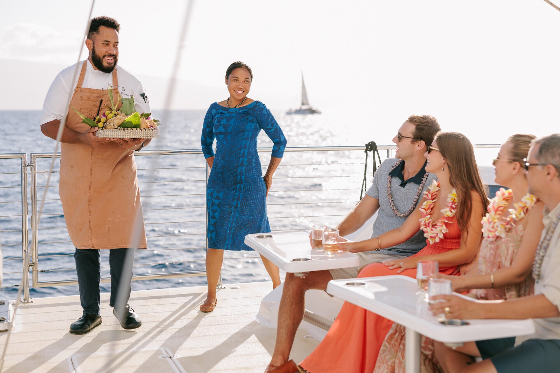 Two employees on the deck of a boat present a platter of fruit to couples sitting in pairs, wearing leis and holding drinks.