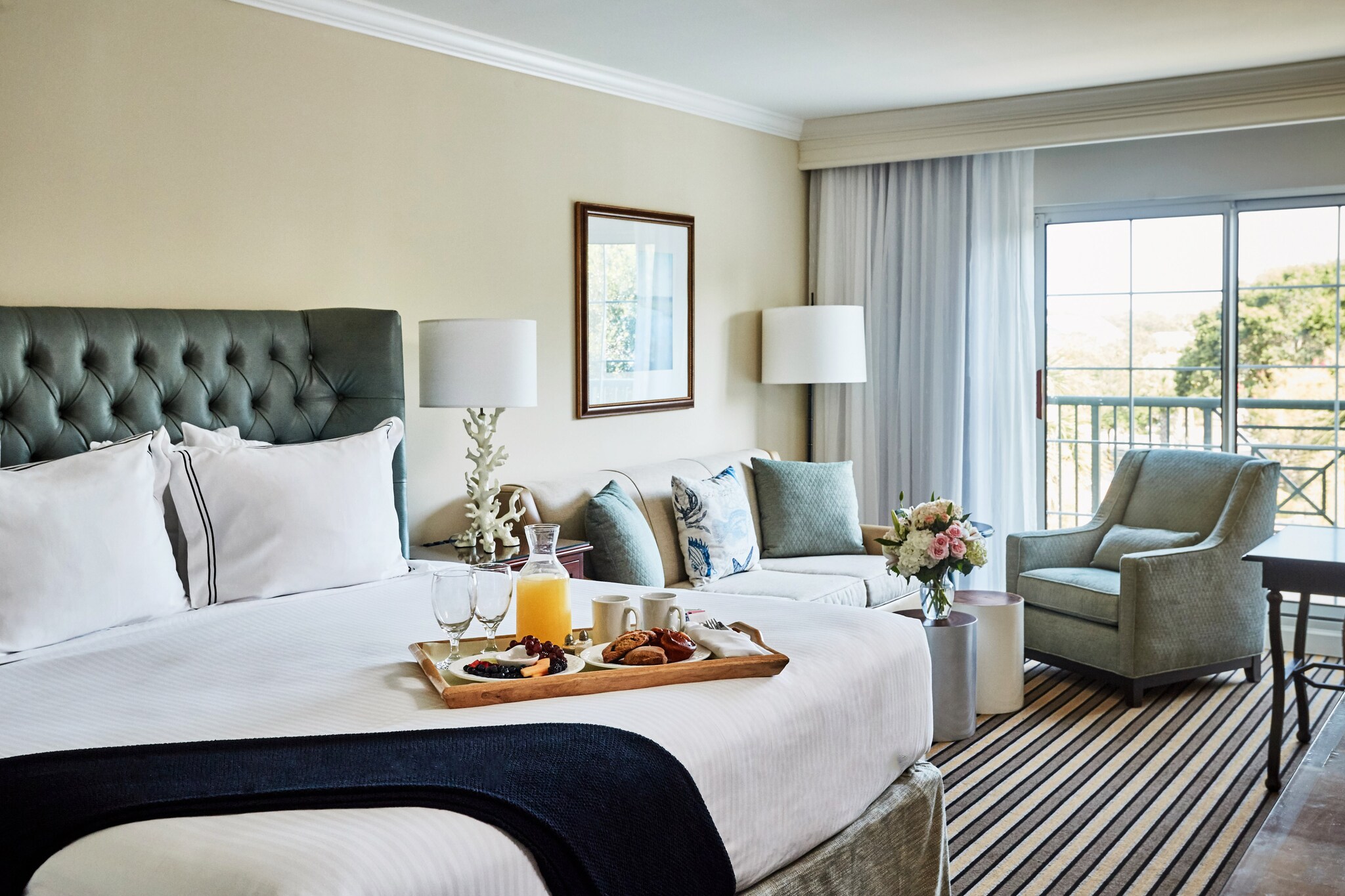 In a sunny guest room, a large bed is laid out with a tray of breakfast, including a carafe of orange juice and a plate of pastries.