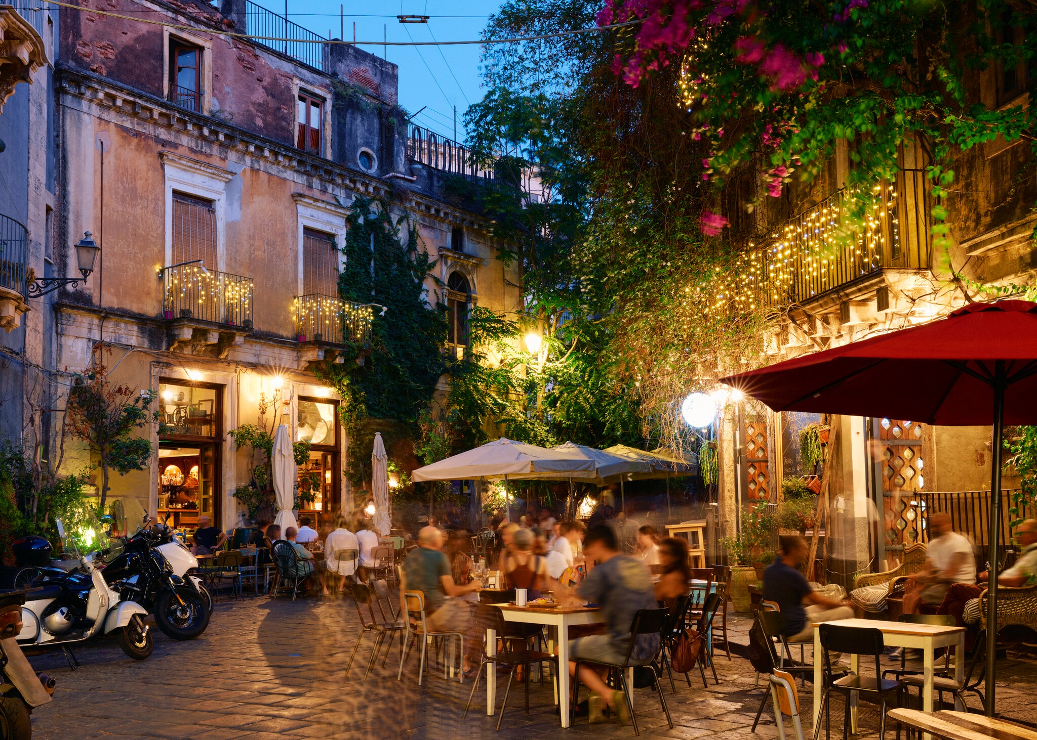 In the evening, a side street is thronged with diners eating at sidewalk tables. Plants and twinkle lights hang from balconies above. 