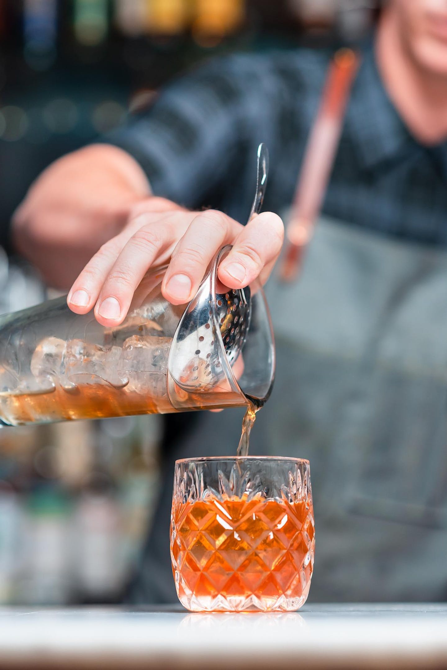Detail shot of a bartender’s hand as they pour an amber mixture through a strainer.