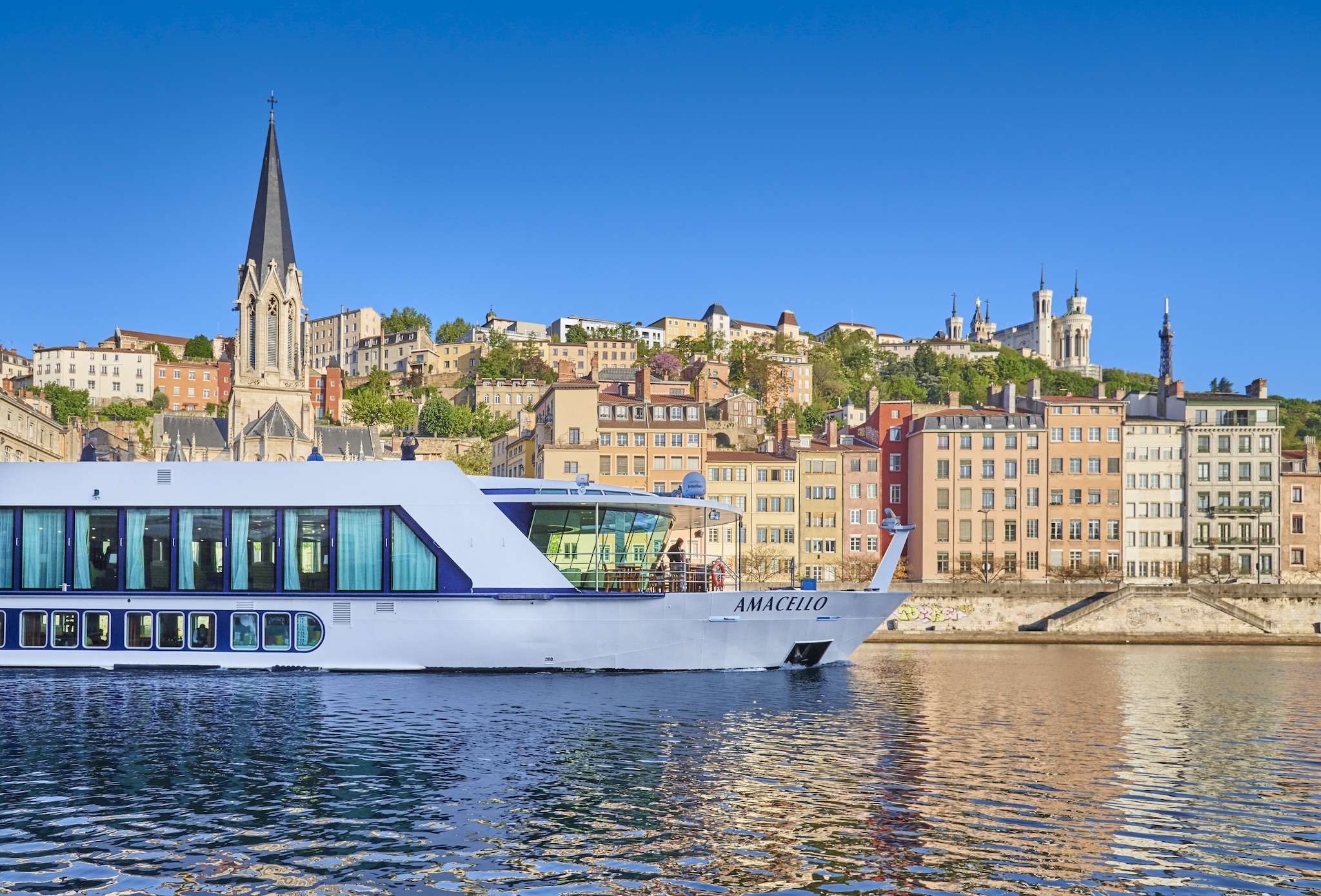 Side view of the long white Amacello ship sailing the Rhône river past the Saint George Church of Lyon on a sunny day.