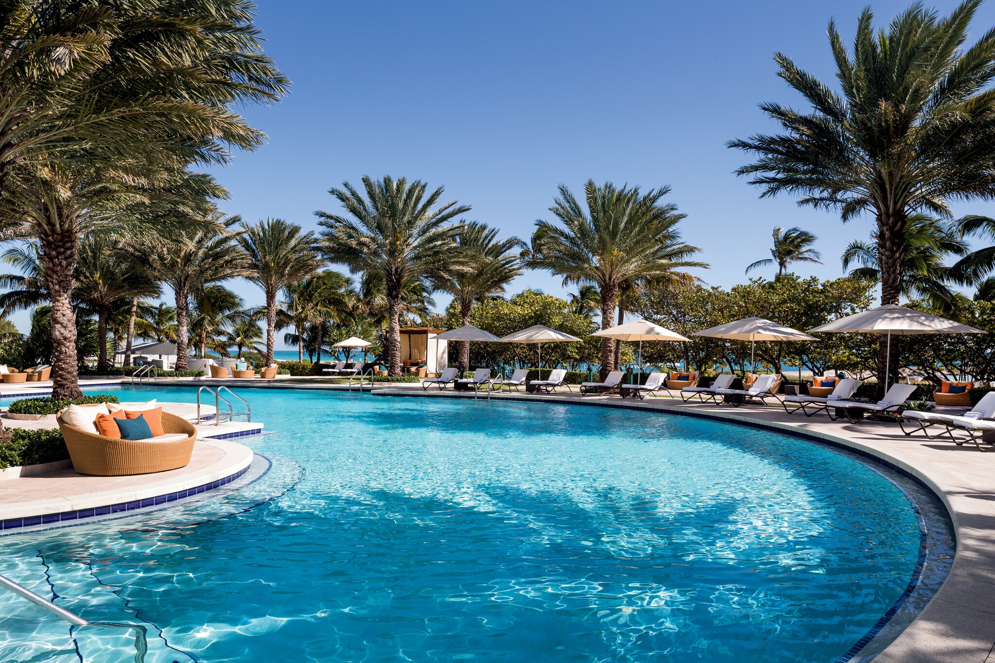 A curved outdoor pool is fringed with seating, umbrellas, and palm trees.