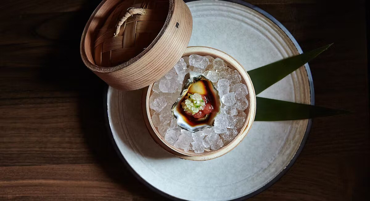 Overhead view of a round plate holding a steamer basket of ice. On the ice is an open oyster holding dark brown liquid and diced vegetables.