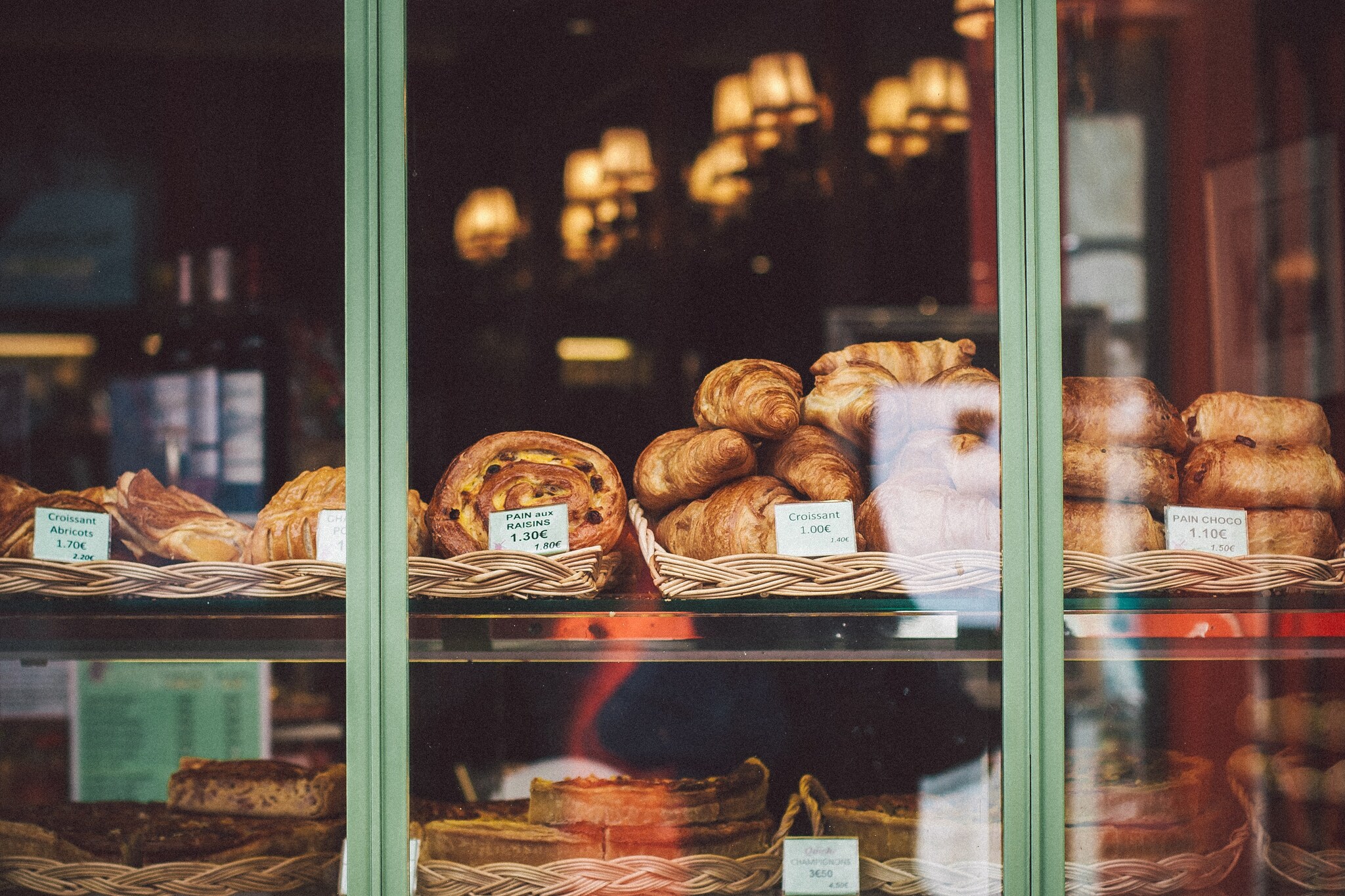 Baskets of plain croissants, along with chocolate, raisin, and apricot varieties, are visible through the windows of a patisserie.