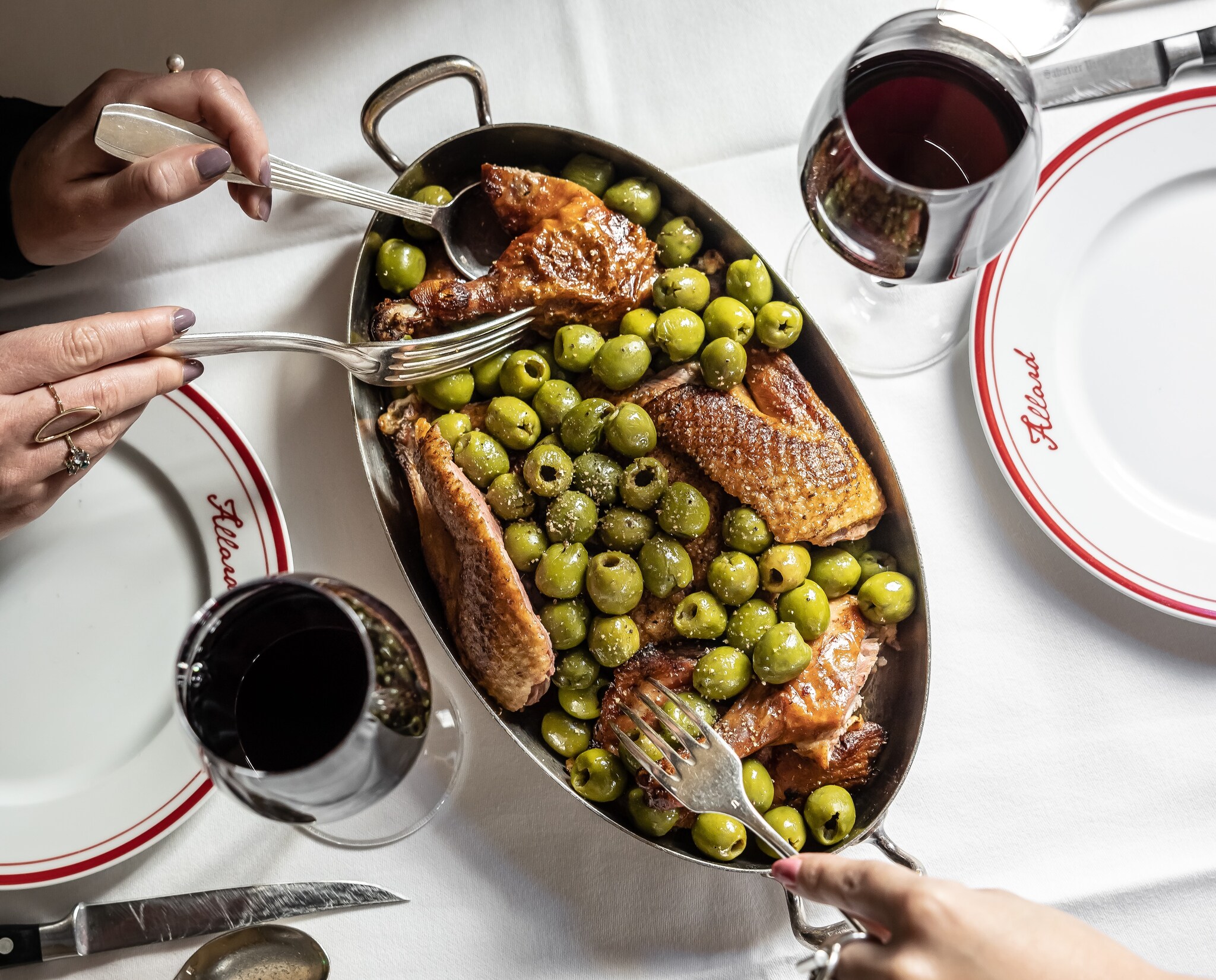 A diner’s hands are visible as they use a spoon and fork to serve themselves from a large dish filled with poultry and green olives.