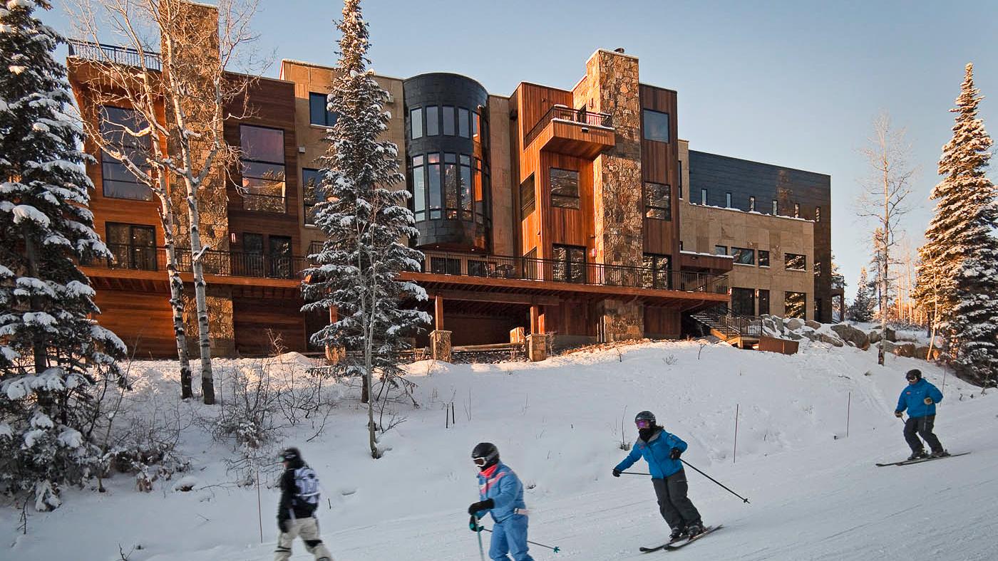 Group of four skiiers going downhill behind large multi-story residential building.