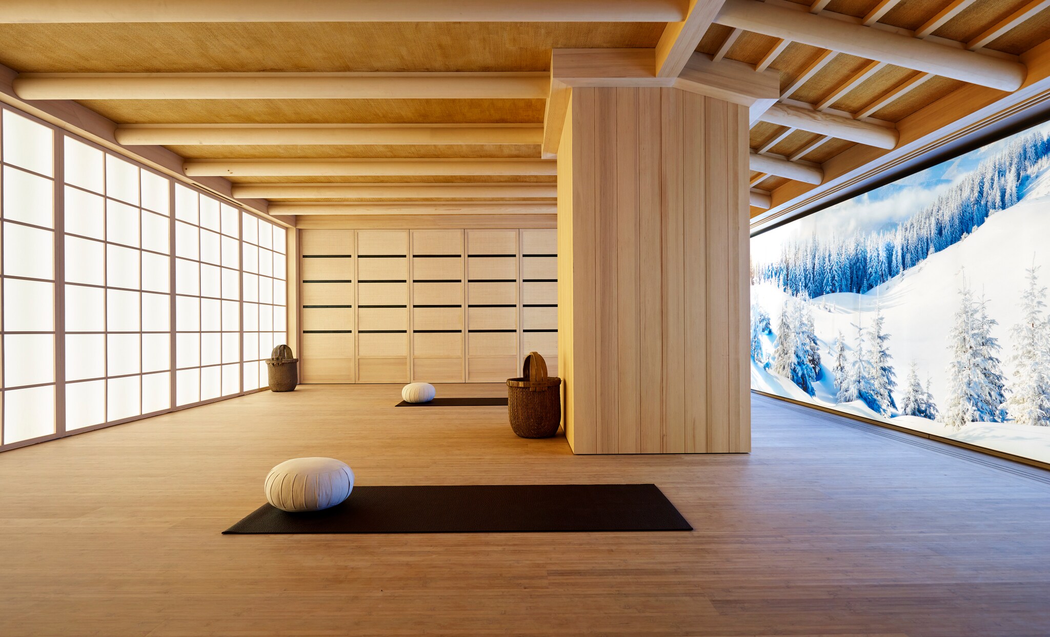 Two black yoga mats with round white pillows in a large, sleek yoga studio. Snowy mountains are visible through a floor-to-ceiling window.