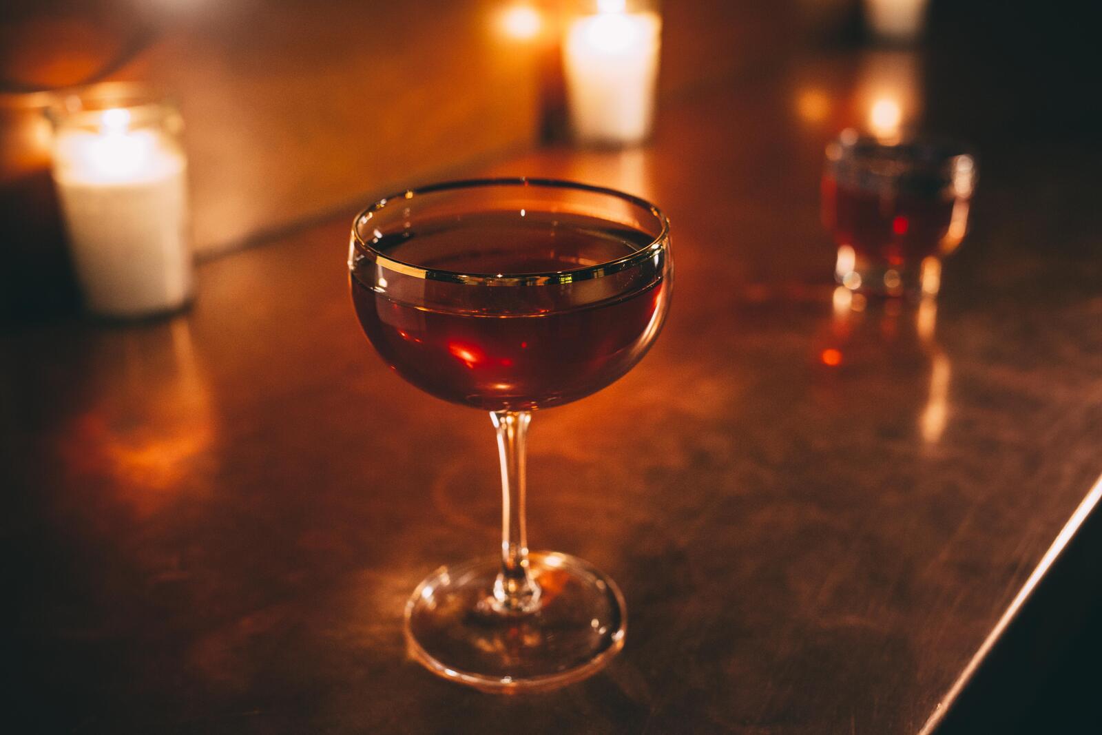 A deep-red cocktail in a gold-rimmed coupe rests on a darkly mirrored bar, with candles and other drinks barely visible in the background.