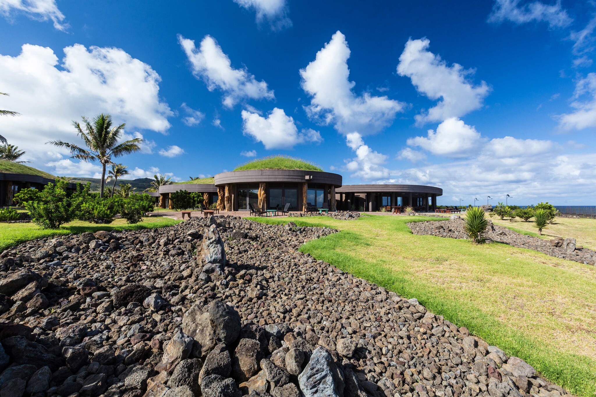 Exterior view of Nayara Hangaroa, a cluster of round buildings with grass domes on top, perched amid grass, bushes, and patches of rocks.