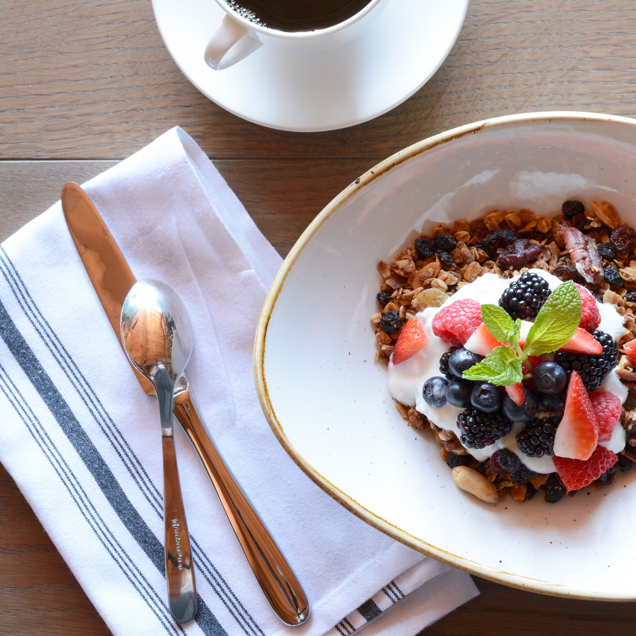 Top-down view of a bowl of granola, yogurt, and berries garnished with mint. Nearby, silverware rests on a blue-striped napkin.