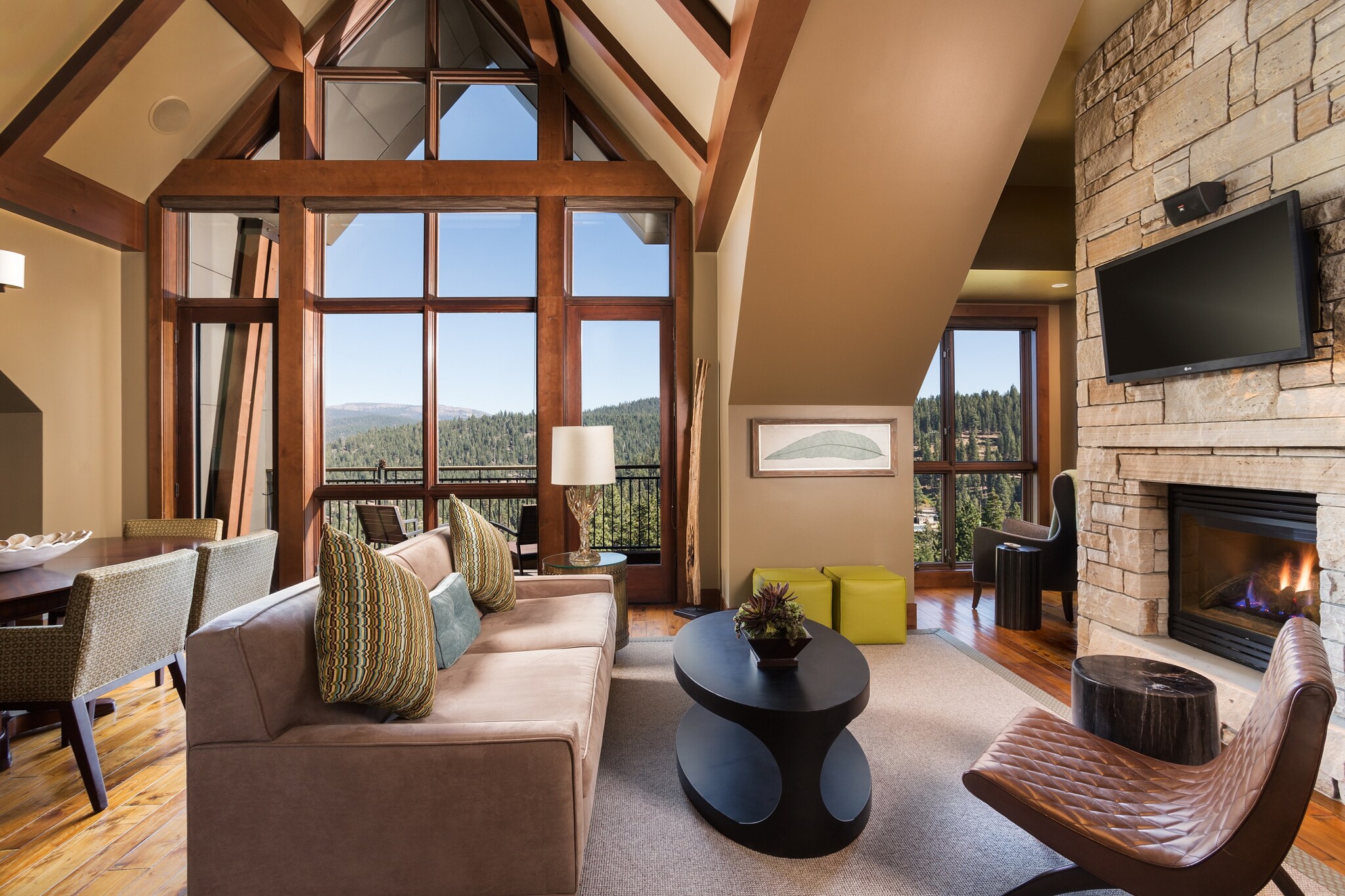 The living area of a three-bedroom residence has a stone fireplace and floor-to-ceiling windows overlooking the forested mountains.