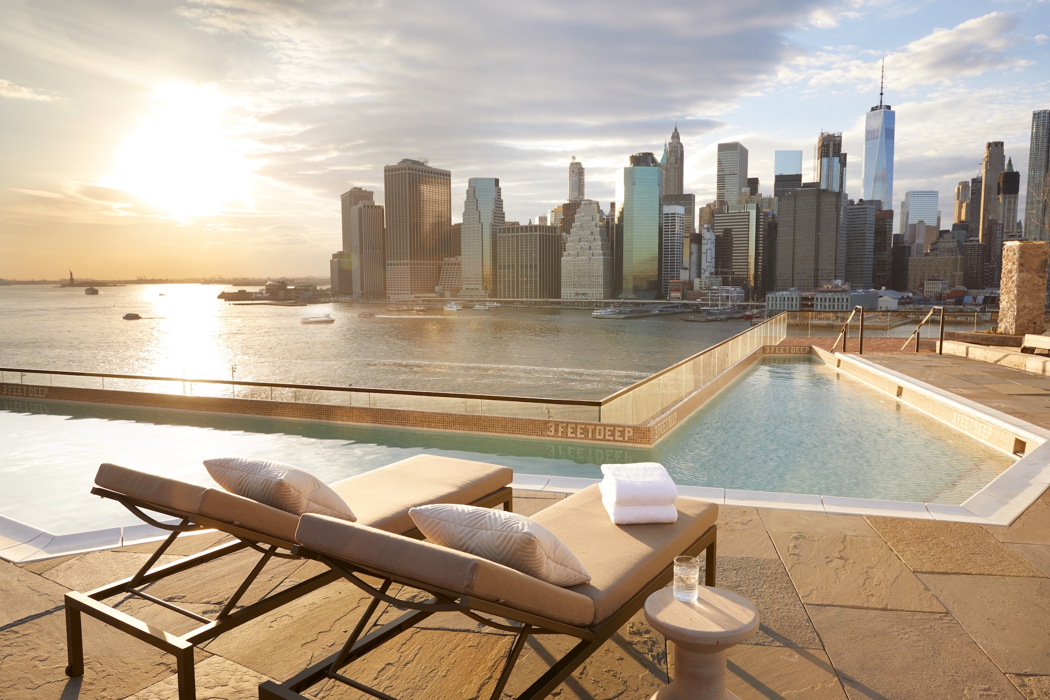 The rooftop pool at 1 Hotel Brooklyn Bridge is irregular, hugging a clear barrier to overlook the water and the Manhattan skyline at sunset.