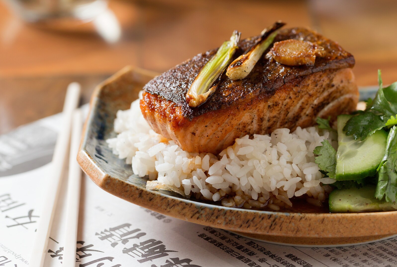 Close-up of glazed salmon garnished with green onion and sitting on a bed of rice. Next to the plate is a Japanese newspaper and chopsticks.