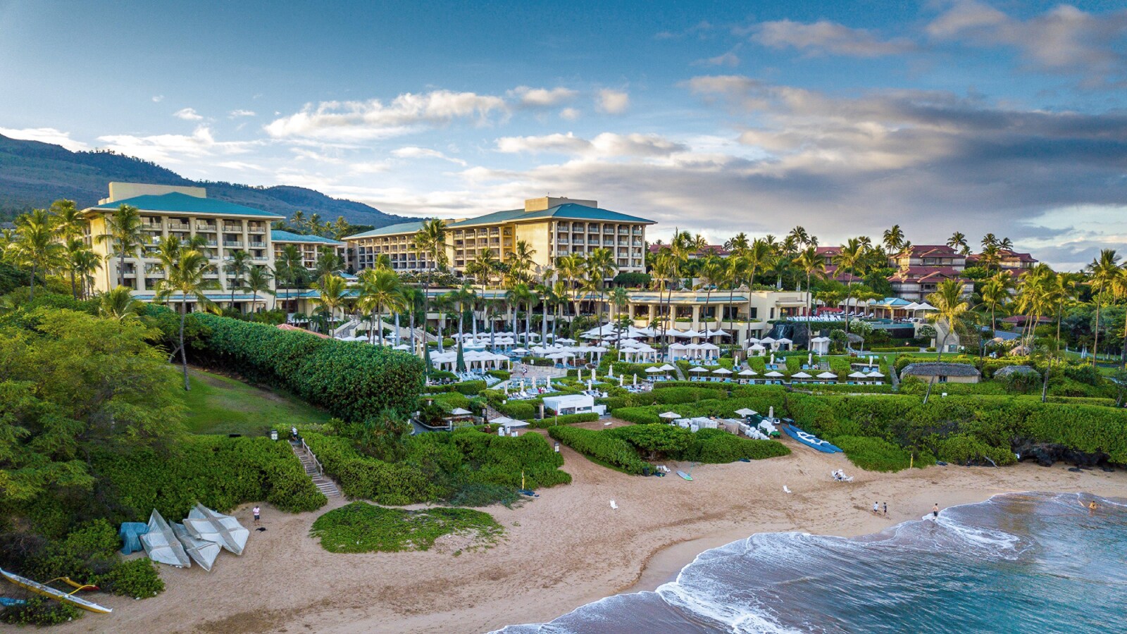 Aerial view from over the water as the ground slopes up from the sandy beach to lush grass and shrubs, through rows of cabanas to the hotel.