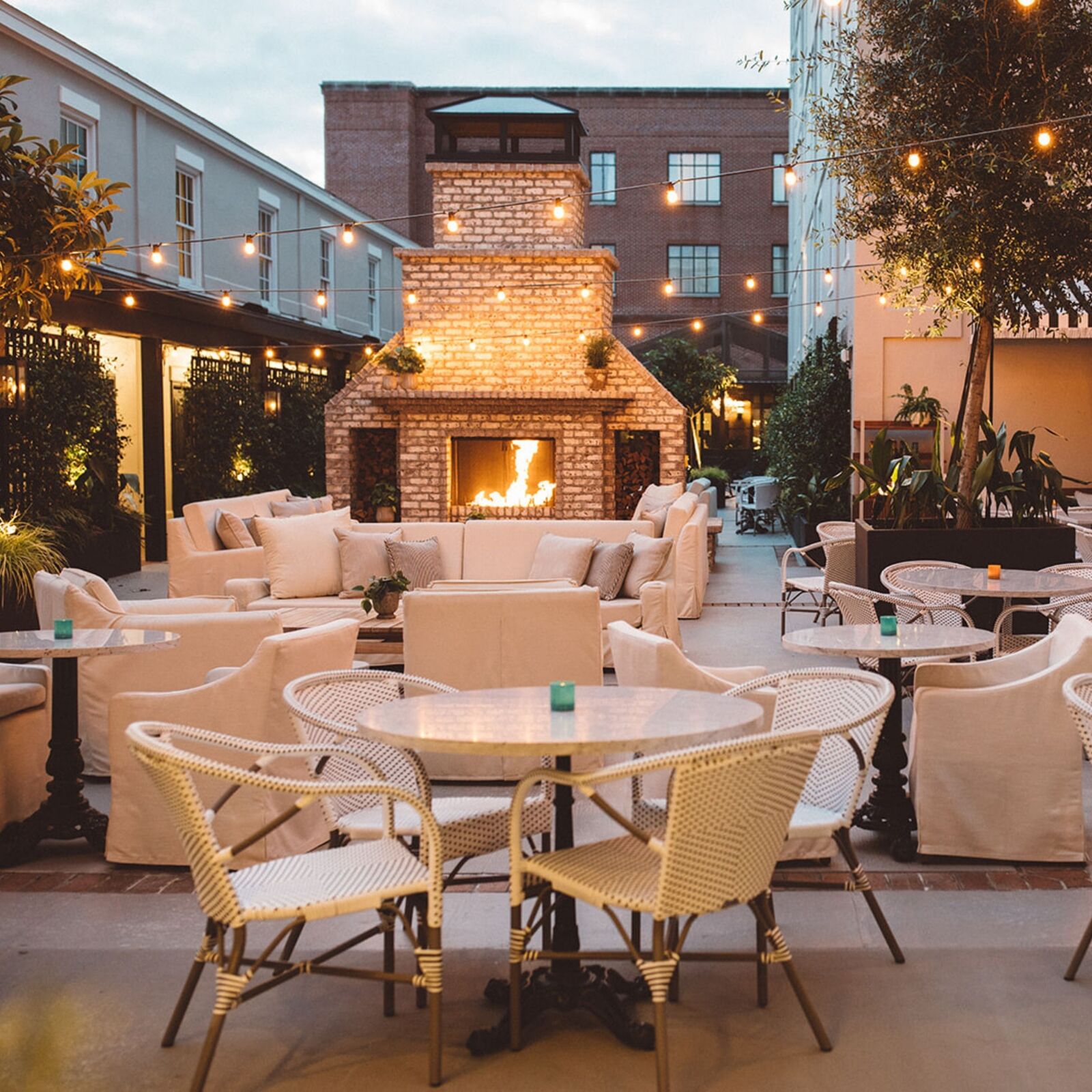 White wicker chairs surround white tables in an outdoor area strung with hanging lights in front of a white brick fireplace.
