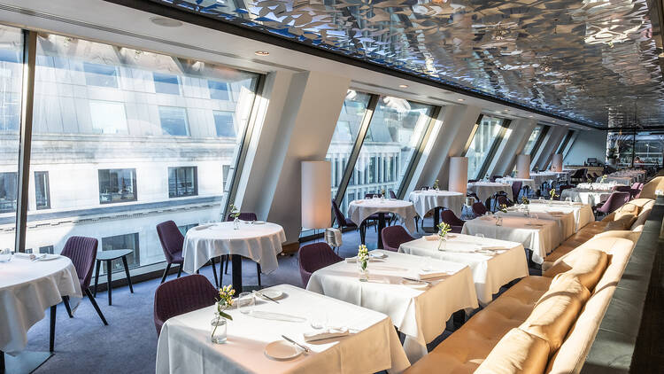The dining room at Angler features rows of small round and square tables under a silver mirrored ceiling, and wall of windows angled in.