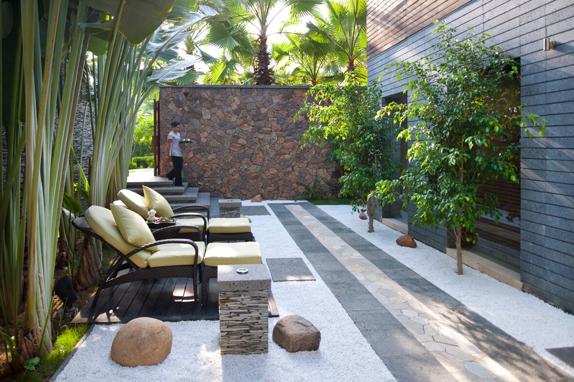 Outside the spa treatment area, a gray path cuts through white gravel, and two black-and-green lounge chairs are arrayed under tall greenery.