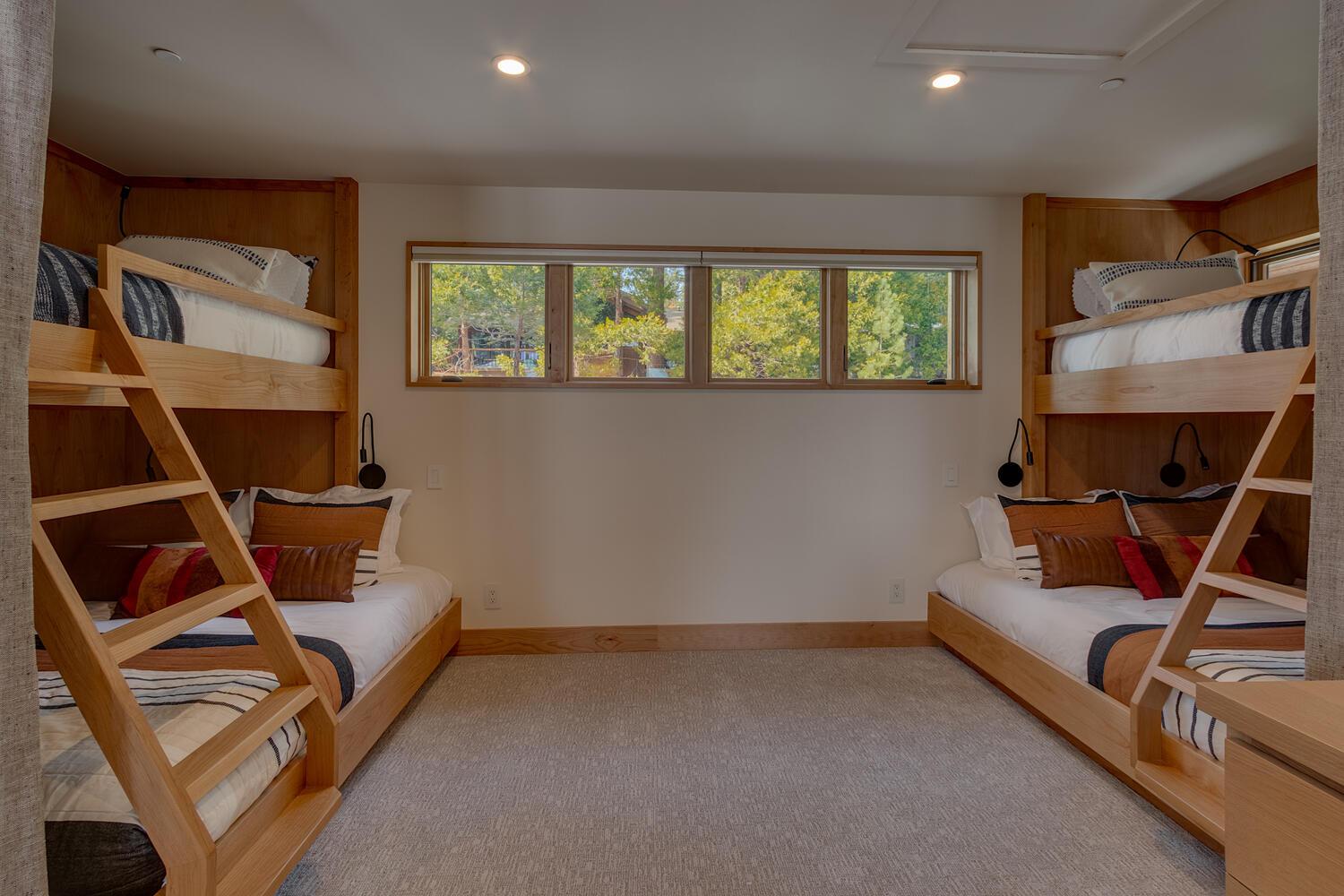 A guest room is furnished with two sets of bunk beds on either side of a set of windows. The pillows and bedspreads on the beds are striped.