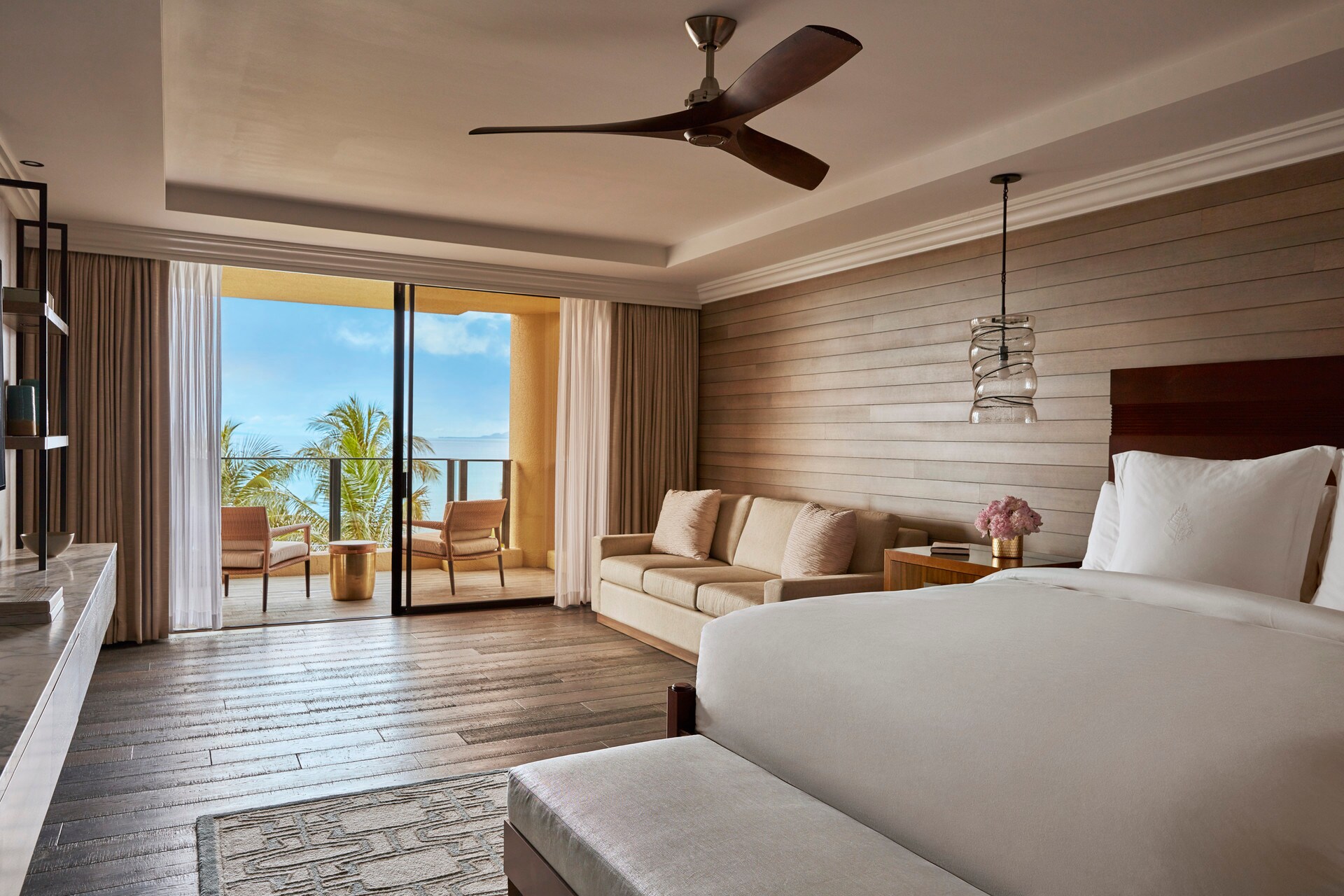 In a guestroom with wood-paneled walls and hardwood floors, gauzy white and taupe curtains frame glass doors to a balcony facing the beach.