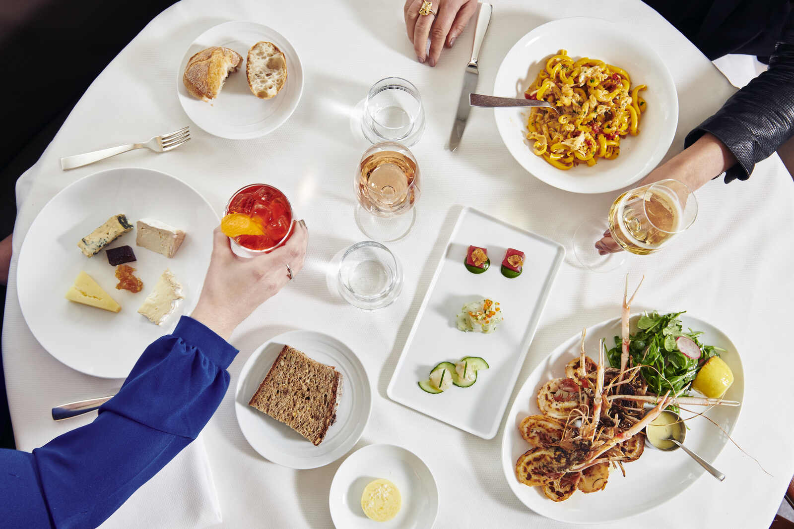 Multiple sets of hands reach across a dining table, holding wine glasses and cocktails over bowls of pasta, bread, cheese, and shellfish.