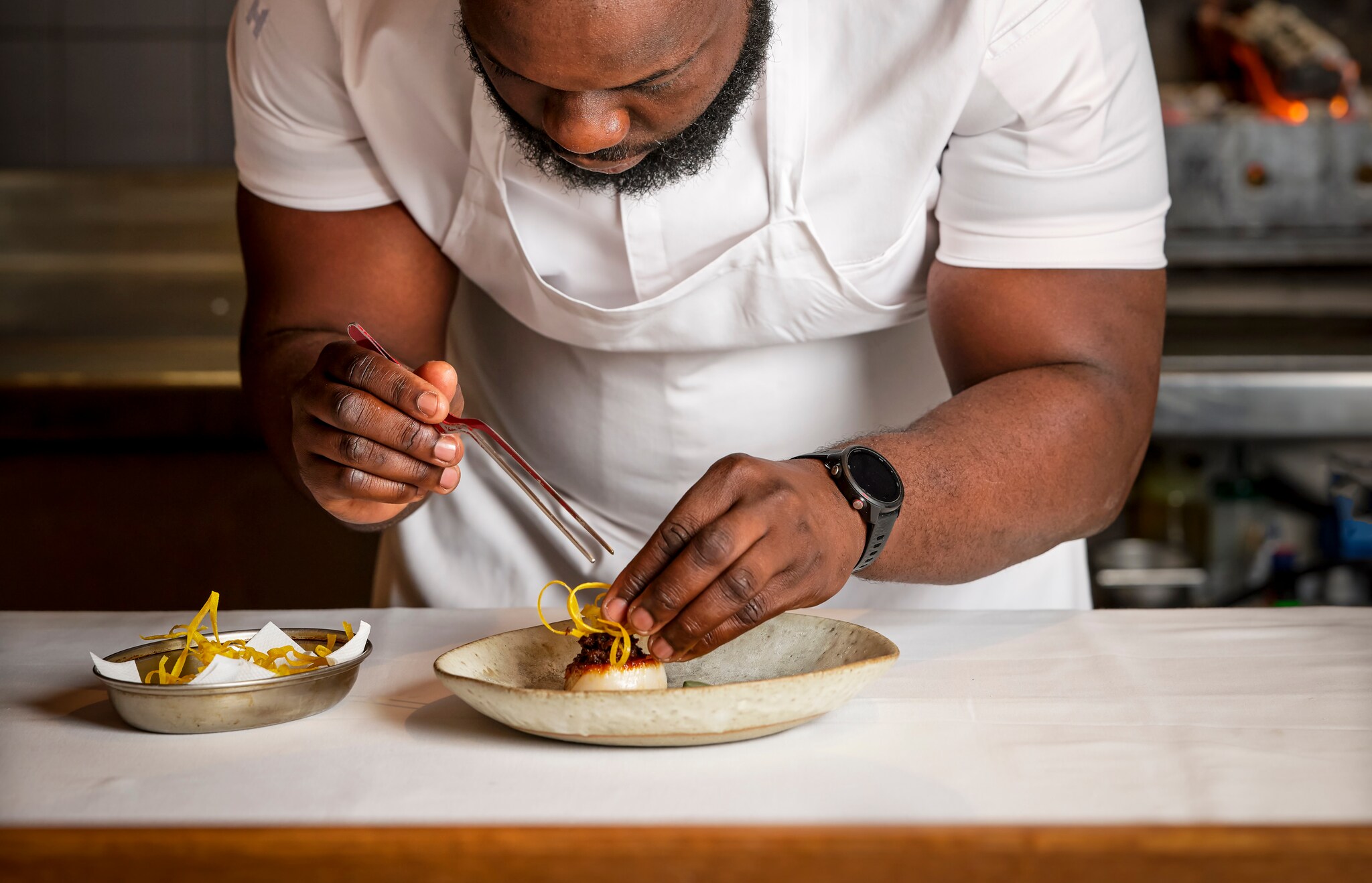 A Black male chef with a short beard uses tweezers to carefully arrange a yellow, noodle-like garnish on a dish.