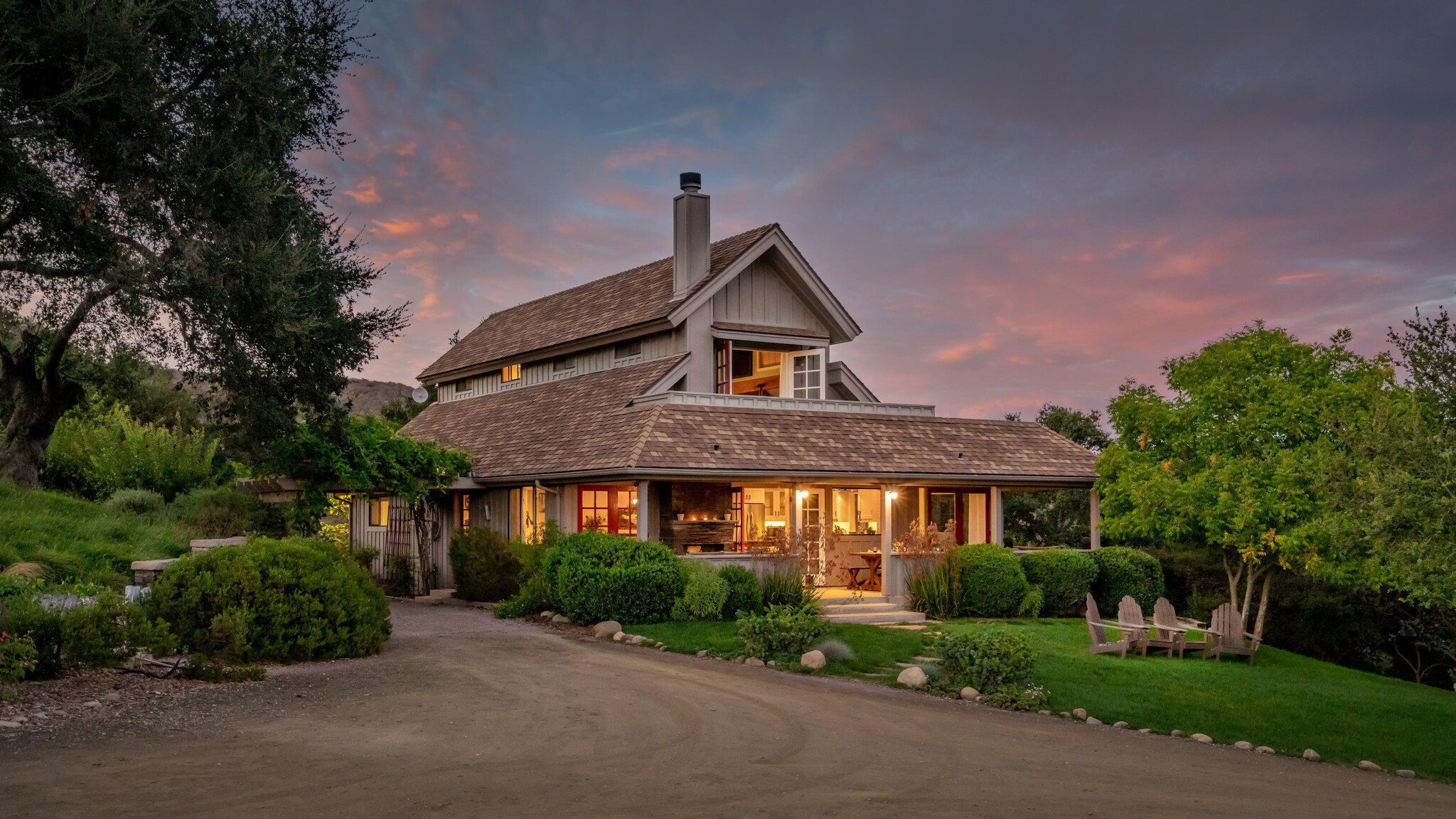 Exterior view of two-story cottage set amongst mature trees and showcasing outdoor seating area in grass.