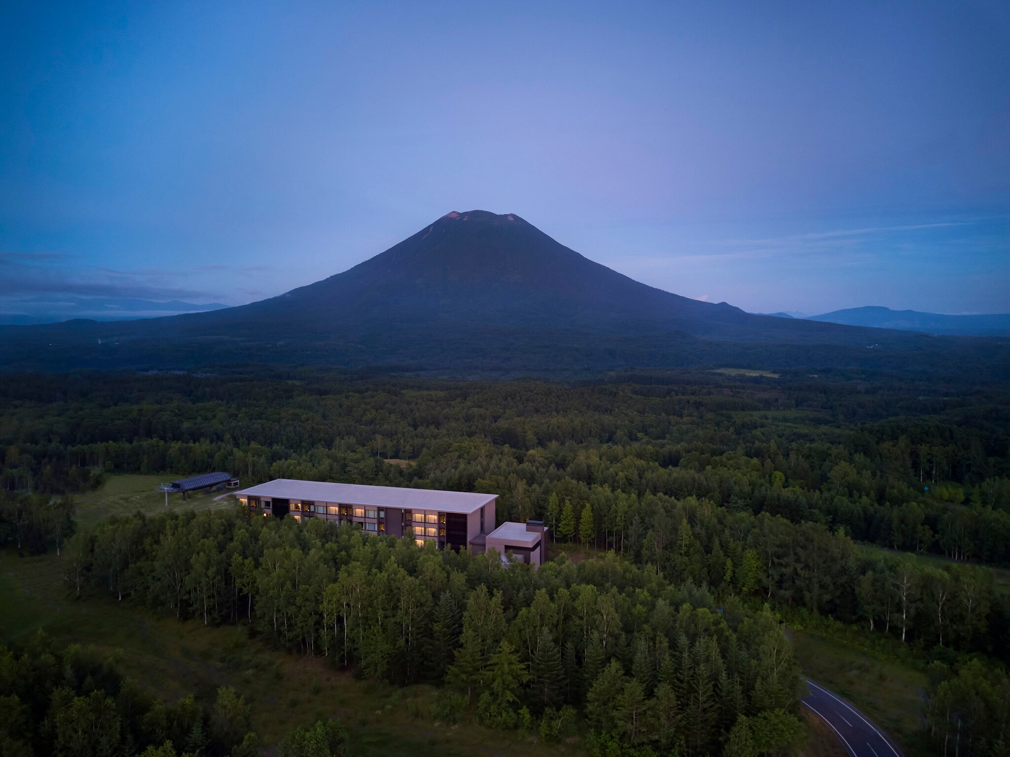 Higashiyama Niseko Village is a pale rectangle surrounded by forest at the foot of a mountain. In the evening, the windows glow golden.