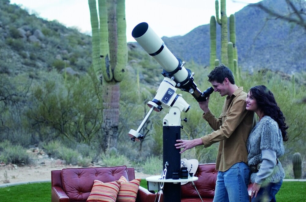 In early evening against the backdrop of the desert, a young couple leans on each other as they look through a telescope.