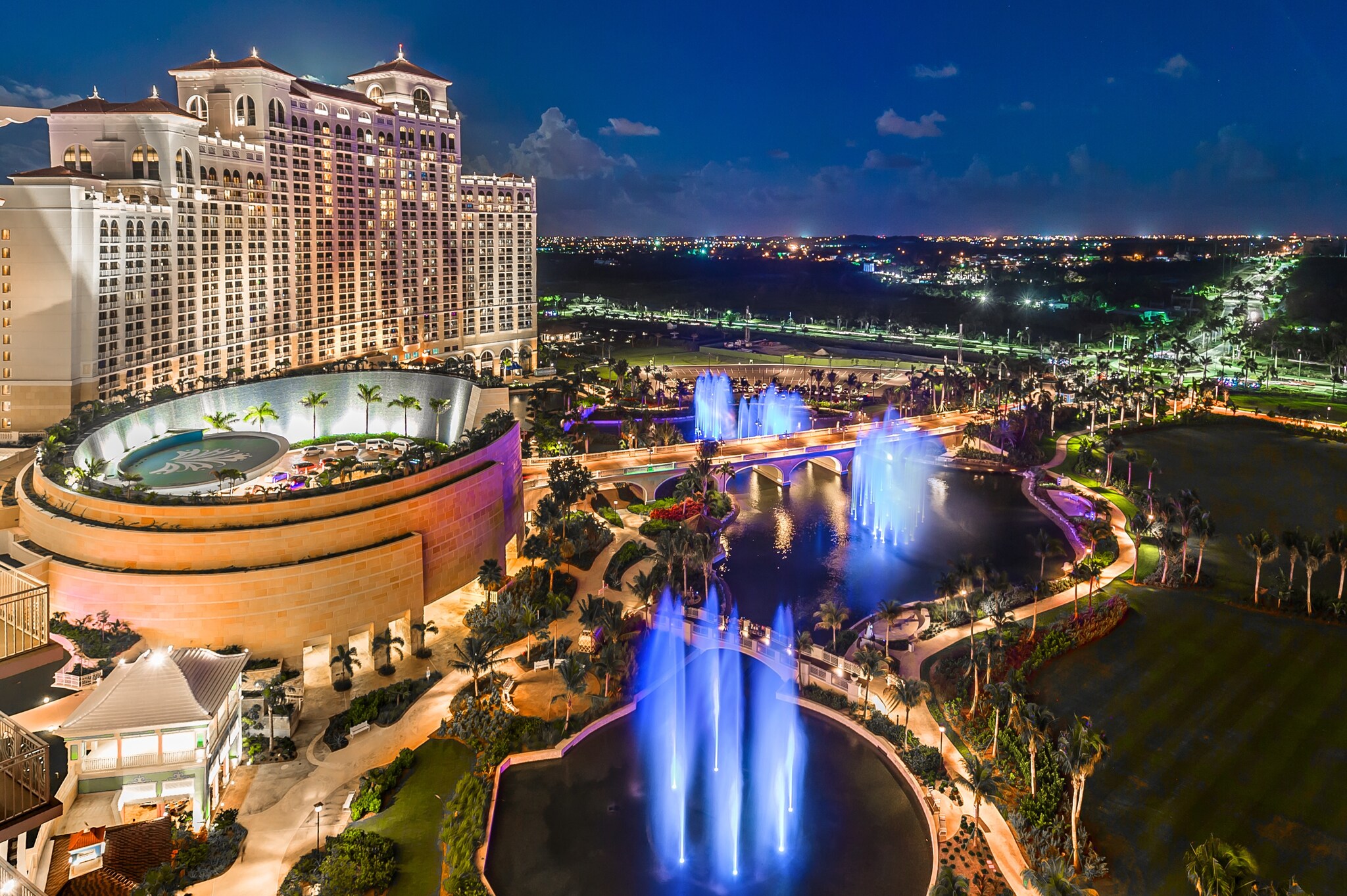 Aerial view of the resort at night. Bright colors light a fountain show around the circular arrival area in front of the grand main building.