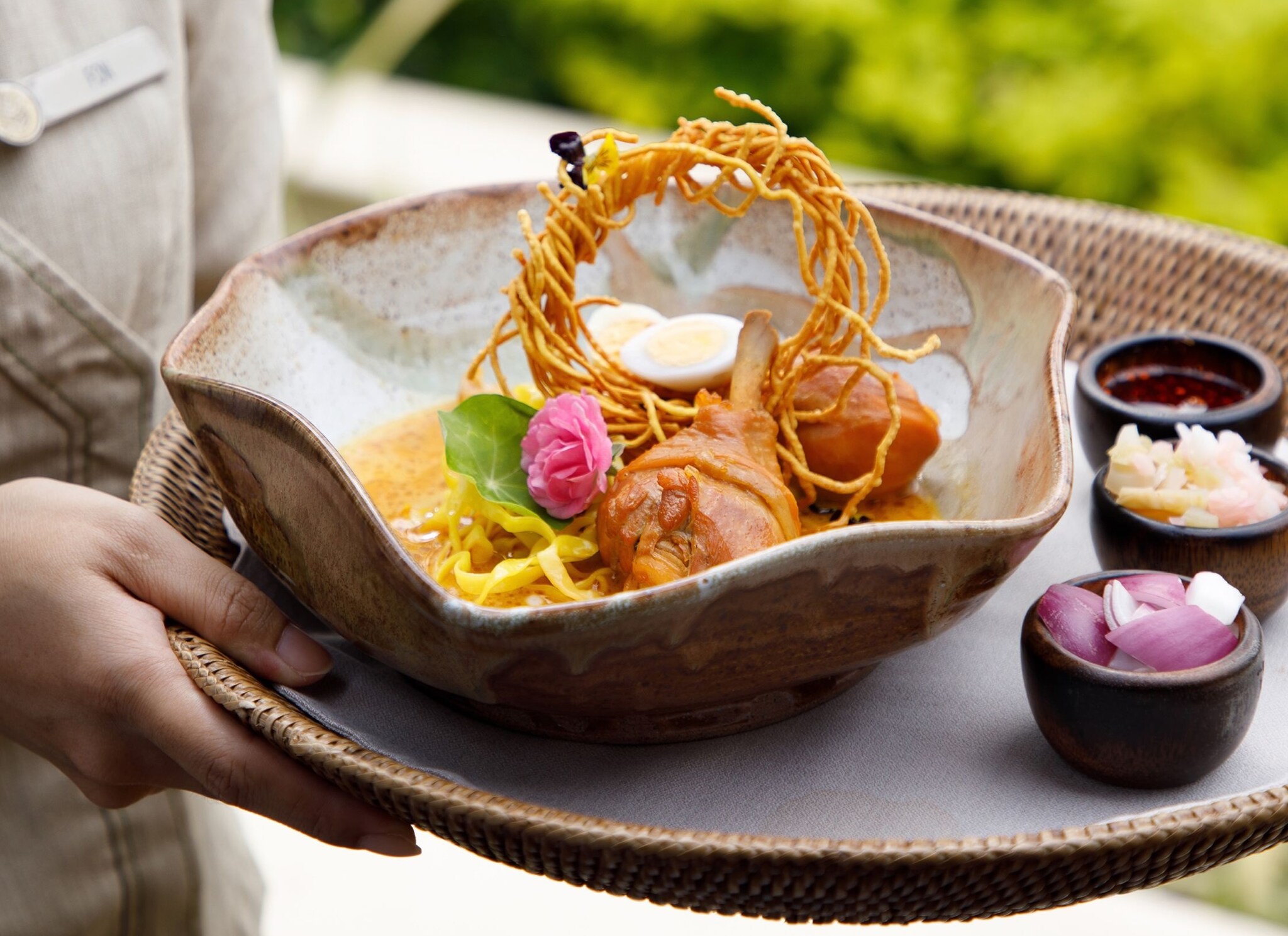 Close-up as a pair of hands carries a tray with a scalloped ceramic dish of curry and noodles, topped with a circle of fried noodles.