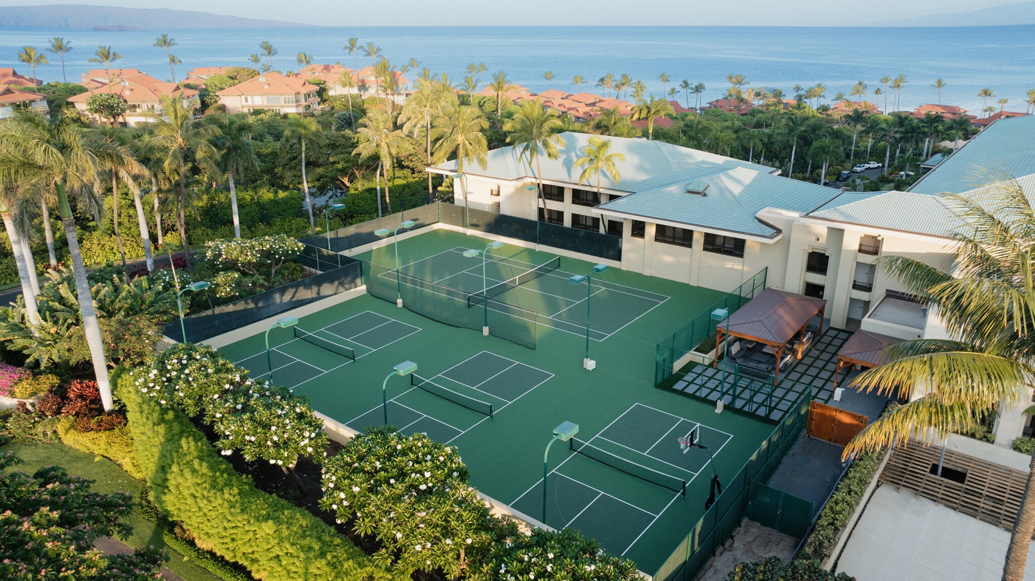 Aerial view of the green tennis courts on a bright morning. The courts are surrounded by thick hedges, palm trees, and low white buildings.