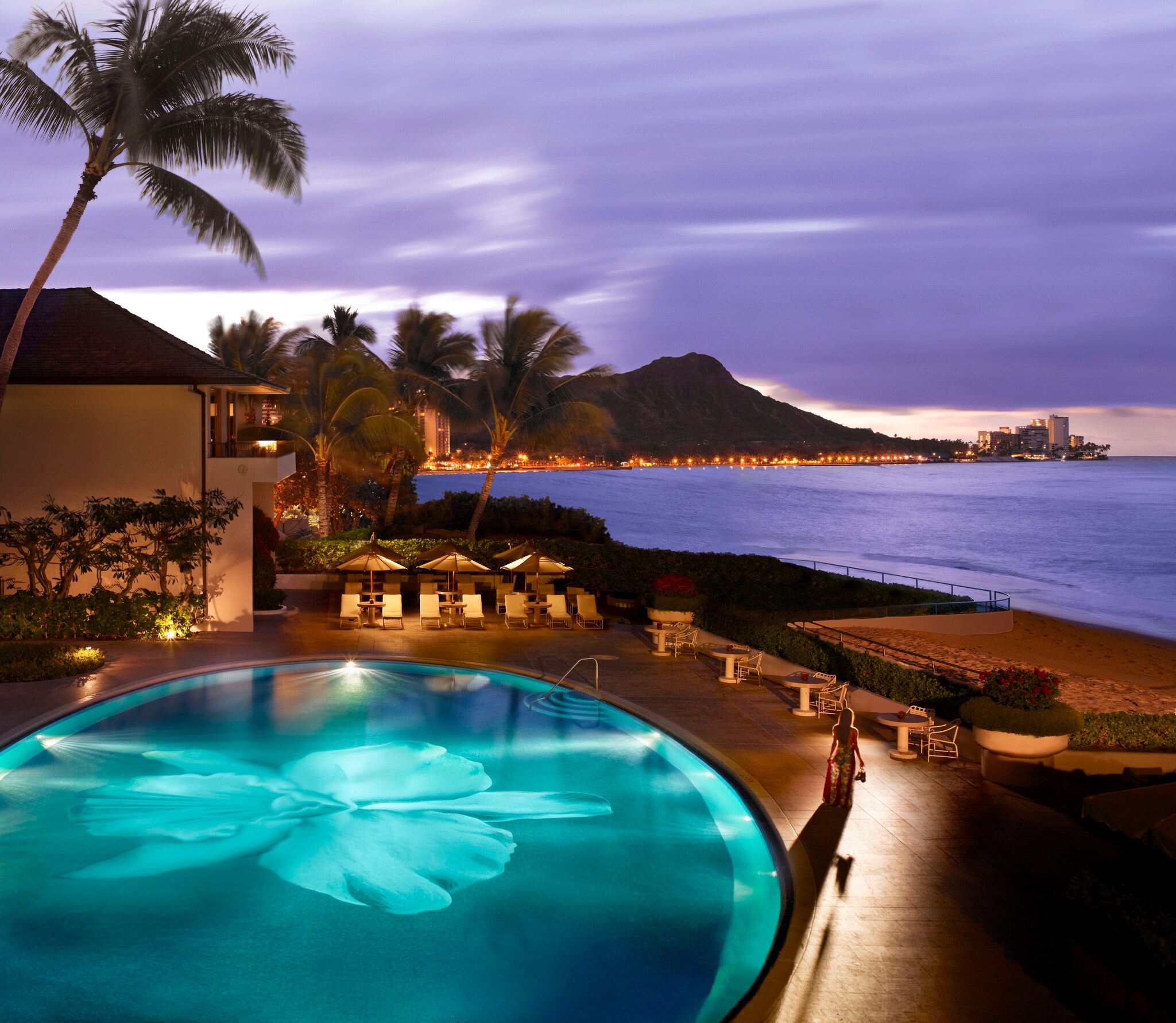 Evening view over the hotel pool as a woman walks towards the beach beyond. Evening makes the clouds and ocean purple.