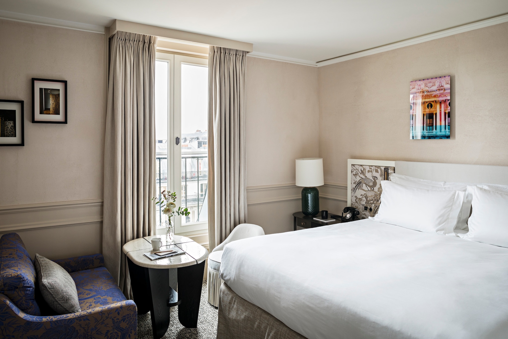 In a room, a big white bed is flanked by a blue-and-bronze armchair and locally-inspired artwork. Glass doors open onto a balcony.