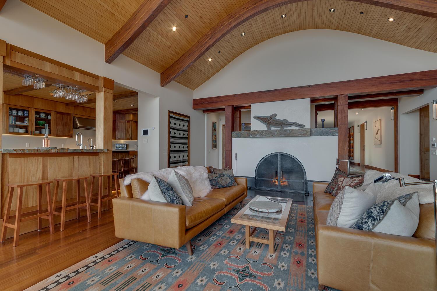 Beyond the tan leather couches of the living room, a fireplace is lit. A bar with stools separates the living room from a kitchen.