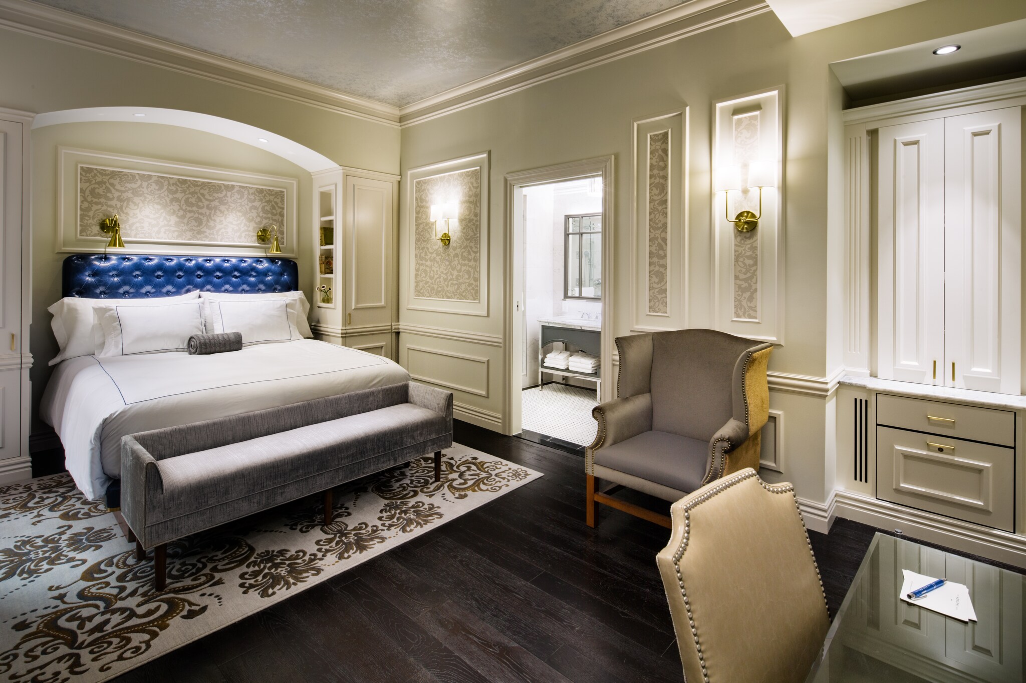 A guest room at The Adelphi Hotel has a desk, an armchair, and a large bed, all decorated in white and gray except for a blue headboard.