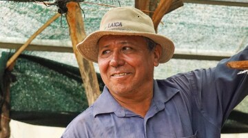 A tanned man wearing a fishing cat and linen shirt looks into the distance while standing under a green canopy.