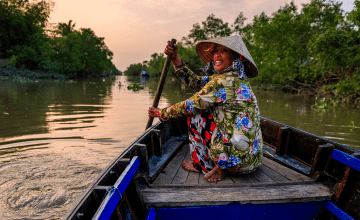 A woman in a conical nón dang hat and brightly patterned clothes paddles a boat down a river, looking back at the camera.