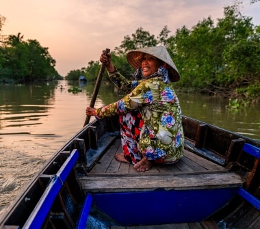 A woman in a conical nón dang hat and brightly patterned clothes paddles a boat down a river, looking back at the camera.