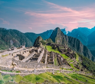 The historic sanctuary of Machu Picchu curves along the top of a grassy mountain, with more peaks rising into pink clouds beyond.