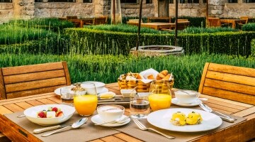 A wooden table and chairs are arrayed outdoors with breakfast, including cut fruit, eggs benedict, and glasses of orange juice.
