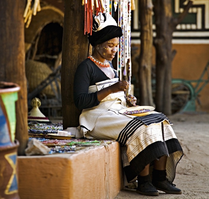 A Xhosa woman wearing a black-and-white patterned wrap and head wrap sits at a market hand-beading jewelry.